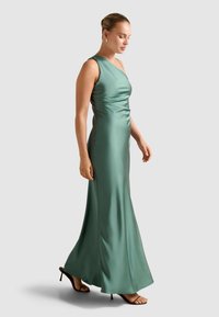 Frau in schulterfreiem, grünem Satin-Evening-Gown mit einem Schulterträger und braunen High Heels, im Profil vor einem einfarbigen Hintergrund stehend.