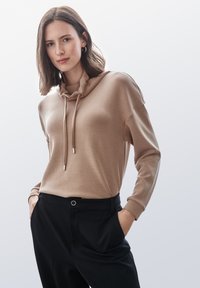 Felpa beige con collo a sciarpa e lacci, abbinata a pantaloni neri. Il tessuto appare liscio, evidenziando una vestibilità comoda.