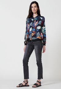 Giacca bomber floreale colorata con sfondo nero, chiusura a zip, polsini a coste, abbinata a jeans neri con borchie e sandali con cinturini.