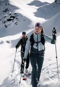 Two individuals trek through snow. The woman wears a light blue and dark gray ski jacket; the man wears a dark green and black jacket.