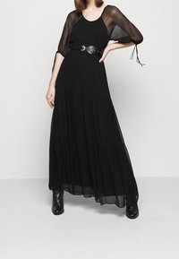 Czarna długa sukienka maxi z przezroczystymi długimi rękawami, teksturowaną tkaniną, warstwowym krojem i szerokim czarnym paskiem. Zestawiona z czarnymi botkami za kostkę.