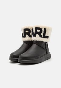 Botas de tobillo de cuero negro con un puño de felpa crema que presenta letras negras en negrita en la parte superior. Textura suave y diseño de punta redondeada.