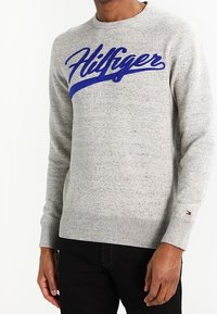 Szary sweter dzianinowy z okrągłym dekoltem, z niebieskim haftowanym logo "Hilfiger" na przodzie. Dodatkowo, ribbowe mankiety i dolny brzeg.