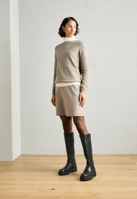 Stivali neri fino al ginocchio con suola spessa, indossati con un maglione e una gonna beige. Texture in pelle liscia e dettagli hardware minimi.