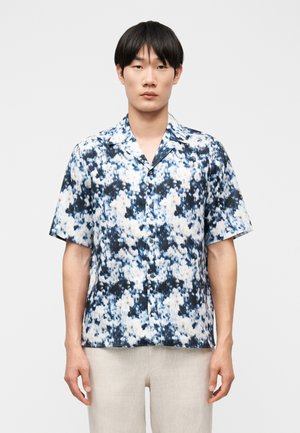 Muž v krátkym rukávom košeli na gombíky s abstraktným kvetinovým vzorom v modrej, bielej a čiernej farbe a svetlými nohavicami na jednoduchom pozadí.