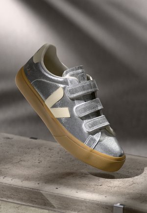 Sneaker sintetico argento con tre cinghie in Velcro, dettagli crema, punta arrotondata e suola in gomma leggermente marrone texturizzata.