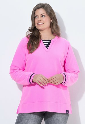 Kvinde med langt brunt hår iført en lyserød sweatshirt over en sort/hvidstribet skjorte, grå bukser, smilende og kigger til siden.