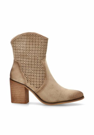 Bota de tobillo beige con parte superior perforada, textura suave de ante, puntera puntiaguda y un tacón de madera grueso. Suela lisa con sutiles acentos.