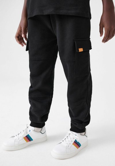 Mustat cargo-housut, jotka on valmistettu pehmeästä kankaasta, sisältävät sivutaskuja ja tekstuuripinnan, yhdistettynä valkoisiin lenkkareihin, joissa on värikkäät raidat.