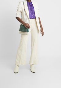 Blazer blanco sobre una blusa púrpura con puntos dorados, combinado con pantalones de campana crema y un bolso cruzado verde. Tejido texturizado y botas de punta.