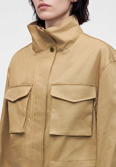 Femme portant une veste beige avec un col montant et deux grandes poches à rabat sur la poitrine.