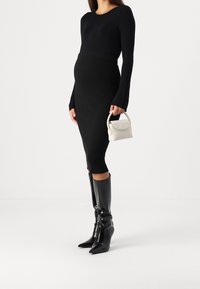 Černé žebrované pletené šaty s dlouhými rukávy, úzký střih, doplněné černými vysokými botami s špičatou špičkou a malou bílou kabelkou.