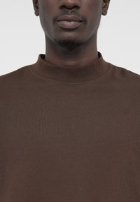 Barna, magas nyakú, hosszú ujjú póló puha anyagból, egyszerű, sima textúrával, minták és látható fémrészletek nélkül.