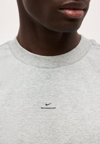 Κοντινό πλάνο ενός ατόμου που φοράει ανοιχτό γκρι μπλουζάκι Nike με την επιγραφή "NIKE STANDARD ISSUE" και το λογότυπο Nike στο στήθος.