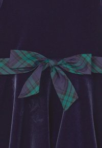 Tmavě modré sametové šaty s širokou kostkovanou mašlí v zelené a tmavě modré barvě uvázanou v pase, která vytváří texturovaný kontrast.