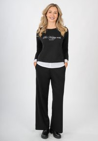 Eine lächelnde Frau mit langen blonden Haaren steht mit den Händen in den Taschen, trägt schwarze Hosen, ein schwarzes Langarmshirt mit der Aufschrift "Be Happy with" und schwarze Stiefel.