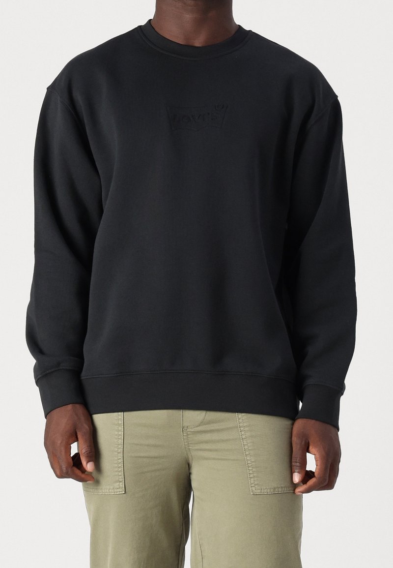 Homme portant un sweat-shirt noir à col rond avec un logo subtil et un pantalon vert olive clair sur un fond uni.