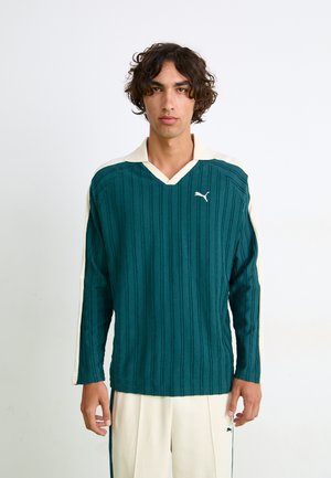 Teal långärmad topp med vertikal ribbning, krämfärgad V-ringad krage och vita sidodetaljer som har en liten vit logotyp på bröstet.