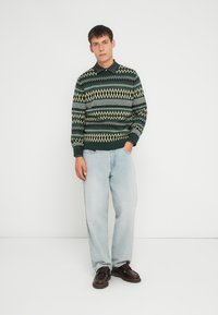 Zelený sveter so vzorom so zúbkami a diamantovými vzormi, skombinovaný so svetlomodrými džínsy a hnedými topánkami. Model stojí na bielom pozadí.