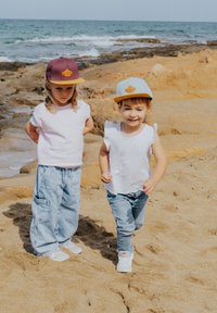 Dos niños en una playa de arena con camisetas blancas. Uno lleva una gorra morada y el otro una gorra azul claro con un logo naranja. Pantalones de mezclilla.