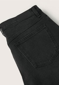 Paire de jeans noirs pliés montrant la poche arrière, le passant de ceinture et une étiquette noire sur la ceinture, sur un fond clair.