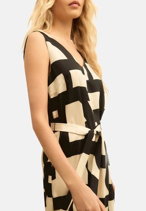 Donna che indossa un vestito smanicato nero e beige con motivo geometrico, scollo a V e cintura annodata in vita, mostrata dalla metà del busto in su.