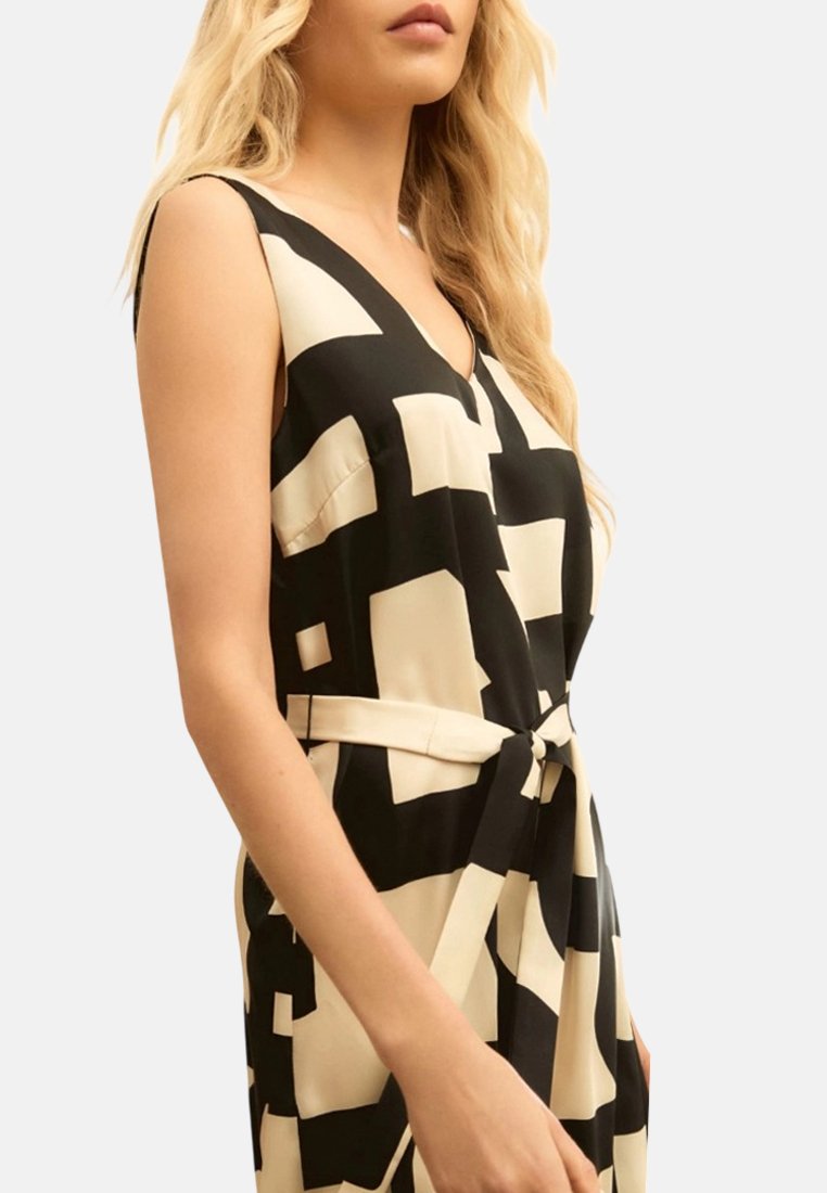 Donna che indossa un vestito smanicato nero e beige con motivo geometrico, scollo a V e cintura annodata in vita, mostrata dalla metà del busto in su.