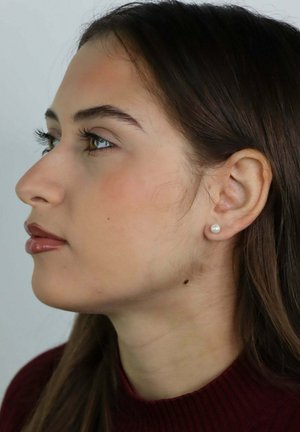 Jonge vrouw met lang bruin haar en parel oorbel, in profiel afgebeeld tegen een effen lichte achtergrond.