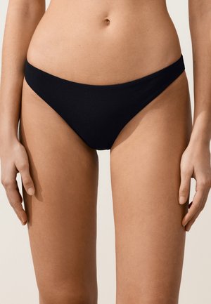 Nő fekete, texturált bikinialsót visel, karjai lazán az oldalán, semleges háttér előtt áll.