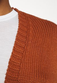 Cardigan en tricot orange rouille à texture côtelée avec un décolleté en V profond, superposé à un t-shirt blanc à col rond.