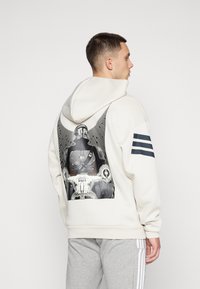 Creme hoodie med en stor grafisk print af en karakter på en motorcykel. Inkluderer tre mørke striber på venstre ærme. Blød stoftekstur.