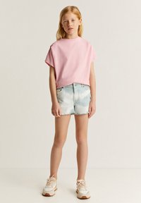 Camiseta de manga corta rosa claro combinada con pantalones cortos de mezclilla azul claro con efecto tie-dye. El atuendo se completa con zapatillas beige con acentos texturizados.