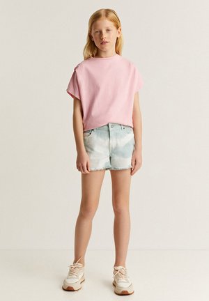 Camiseta de manga corta rosa claro combinada con pantalones cortos de mezclilla azul claro con efecto tie-dye. El atuendo se completa con zapatillas beige con acentos texturizados.