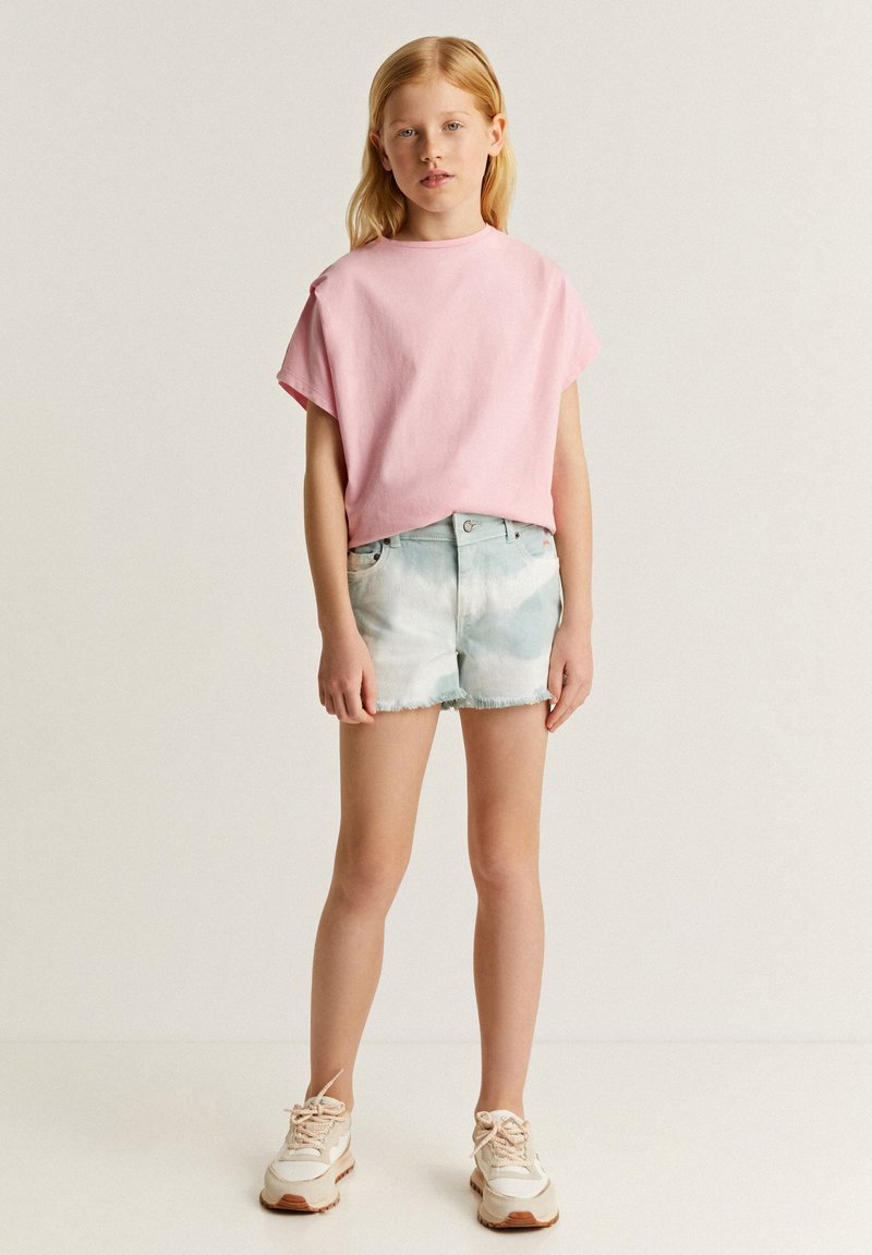 Camiseta de manga corta rosa claro combinada con pantalones cortos de mezclilla azul claro con efecto tie-dye. El atuendo se completa con zapatillas beige con acentos texturizados.
