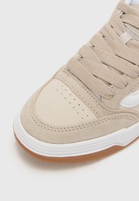 Beige semsketneakers med perforert tå, hvite detaljer og flate lisser. Har en gummisåle og teksturerte stoffdetaljer.