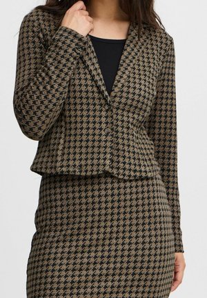 Vrouw draagt een blazer en bijpassende rok met bruin-zwart ruitpatroon over een zwarte top, tegen een effen achtergrond.