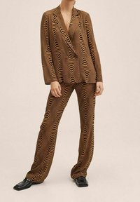 Kobieta ubrana w brązowy i czarny garnitur w geometryczny wzór z rozszerzanymi spodniami i czarnymi butami na kwadratowym obcasie na jednolitym tle.