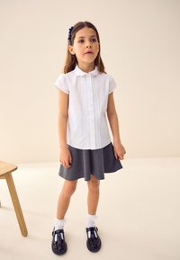 Jeune fille debout portant un chemisier blanc à manches courtes, une jupe grise, des chaussettes blanches à volants, des chaussures noires et une pince à cheveux foncée dans une pièce neutre.