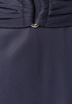 Tamnoplava plisirana tkanina skupljena malim srebrnim gumbom u sredini, pokazujući teksturu i šavove.