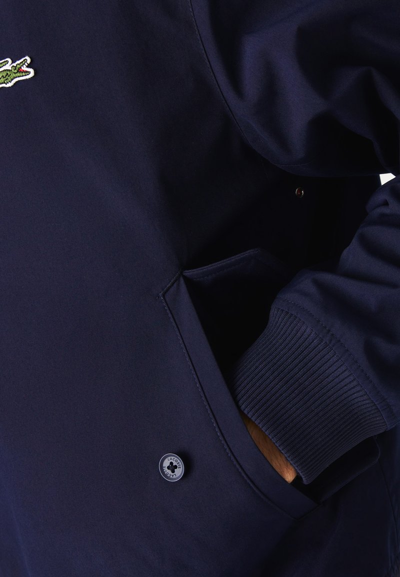 Veste marine avec des poignets côtelés, dotée d'une poche latérale et d'un accent logo rond. Texture lisse et design classique.