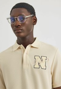 Beige Poloshirt aus strukturiertem Stoff, mit einem auffälligen, gestickten schwarzen "N"-Logo und einem stilvollen Kragen. Die Person trägt eine klare Brille.