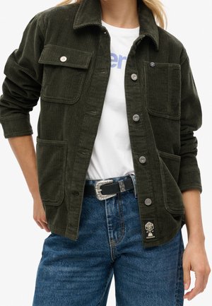 Donkergroene corduroy jas met knopen en voorzakken, gedragen over een wit T-shirt met print en blauwe spijkerbroek met zwarte riem.