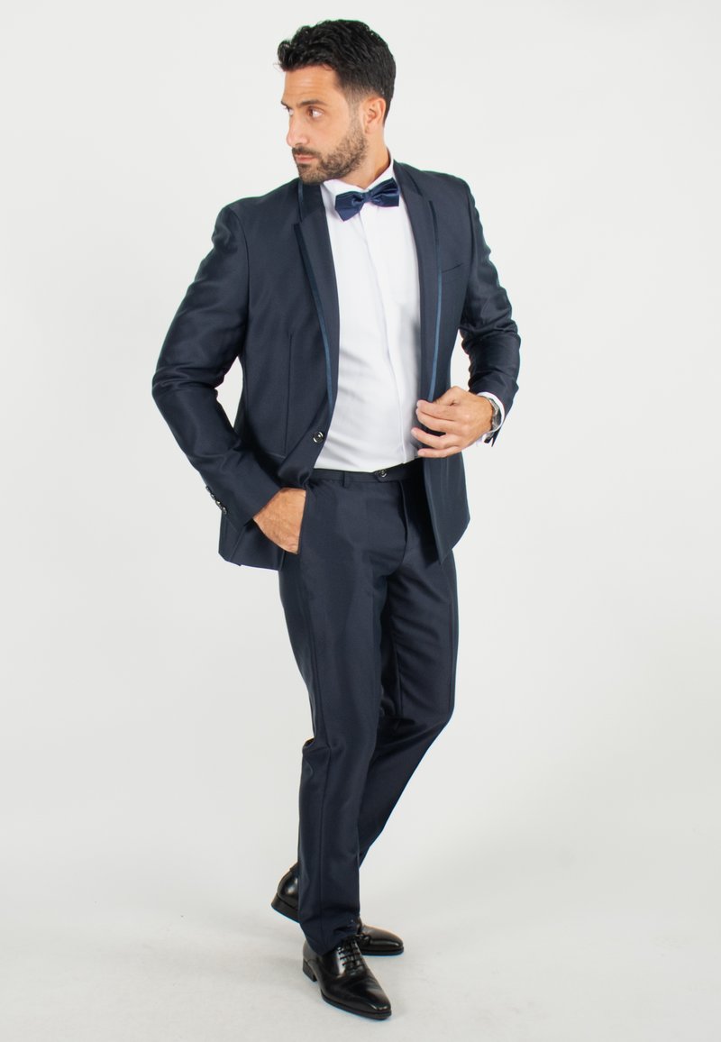 Costume marine avec une finition texturée, chemise blanche et nœud papillon bleu. Coupe ajustée, boutons à l'avant et chaussures habillées noires. Fond simple.