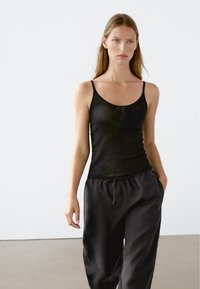 Schwarzes geripptes Tanktop mit dünnen Trägern, mit einem floralen Muster auf der linken Seite. Kombiniert mit lockeren schwarzen Hosen im lässigen Stil.