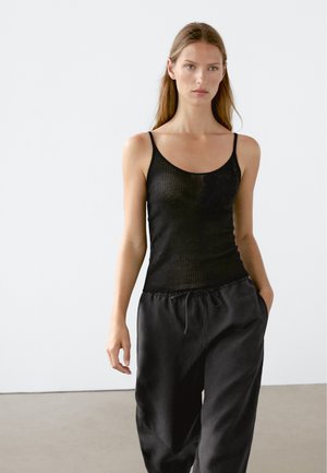 Schwarzes geripptes Tanktop mit dünnen Trägern, mit einem floralen Muster auf der linken Seite. Kombiniert mit lockeren schwarzen Hosen im lässigen Stil.