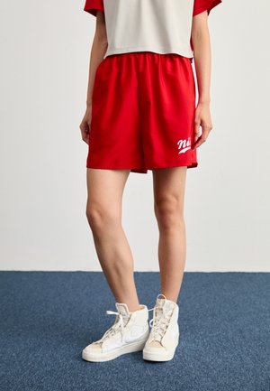 Piros sportnadrág elasztikus derékpánttal és fehér logóval, magas szárú fehér sportcipőkkel párosítva. Az outfitt egy bő szabású szürke felsőt tartalmaz.