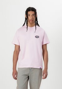Camiseta de algodón rosa clara con mangas cortas, cuello redondo y un pequeño logotipo ovalado negro en el pecho, combinada con unos jeans de color claro.