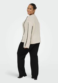 Pull en maille beige avec des accents tressés noirs sur les manches, associé à un pantalon noir ample. Texture lisse et coupe décontractée.