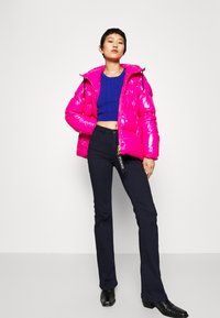 Lys rosa pufferjakke med høyglansf finish, kort design, kombinert med en blå strikket topp og svarte utsvingte bukser.