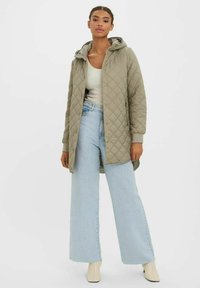 Ljus olivgrön quiltad lång jacka med huva, utrustad med tryckknappar, sidfickor och en avslappnad passform, tillsammans med högmidjade ljusblå jeans.